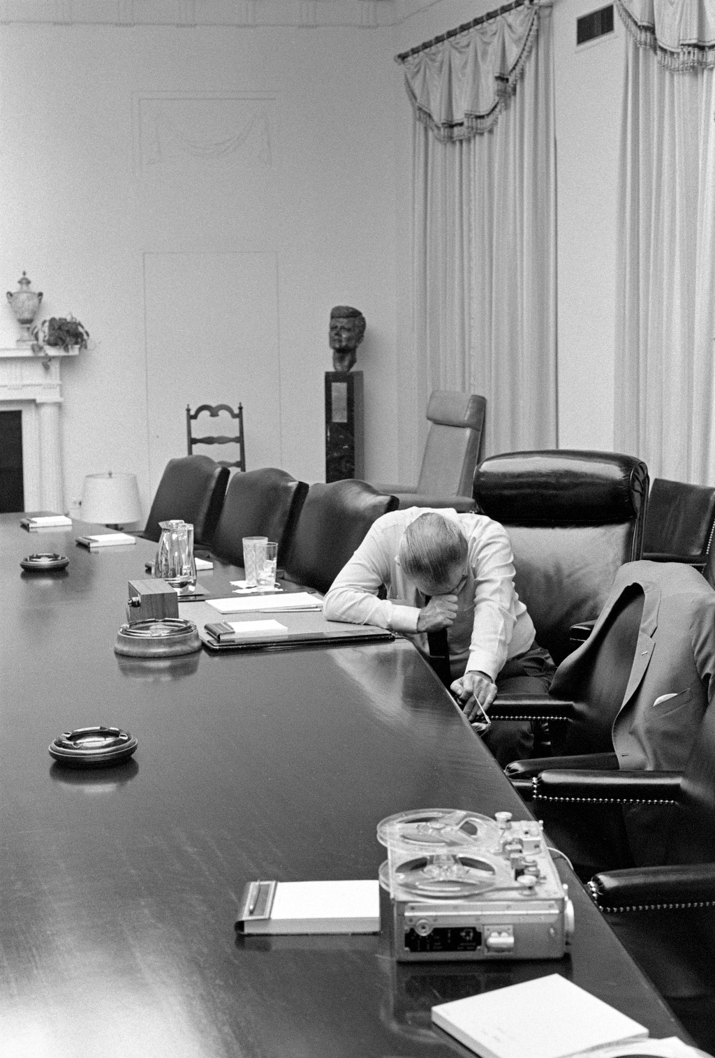 President LBJ seated at the cabinet table, deep in thought with a closed fist to his face. He holds glasses in his other hand, attentively listening to a device on the table.